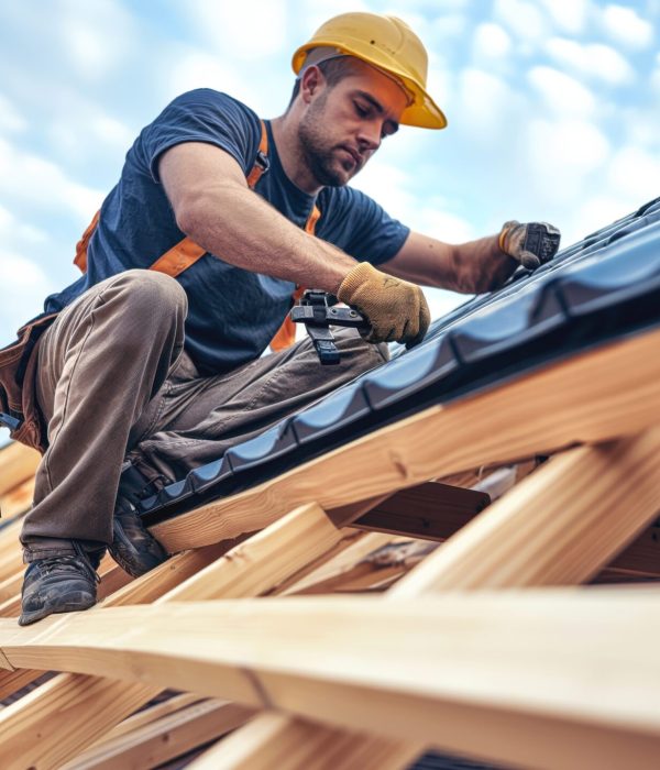 Construction worker wearing a hard hat and safety gear installing roofing materials on a wooden roof framework.